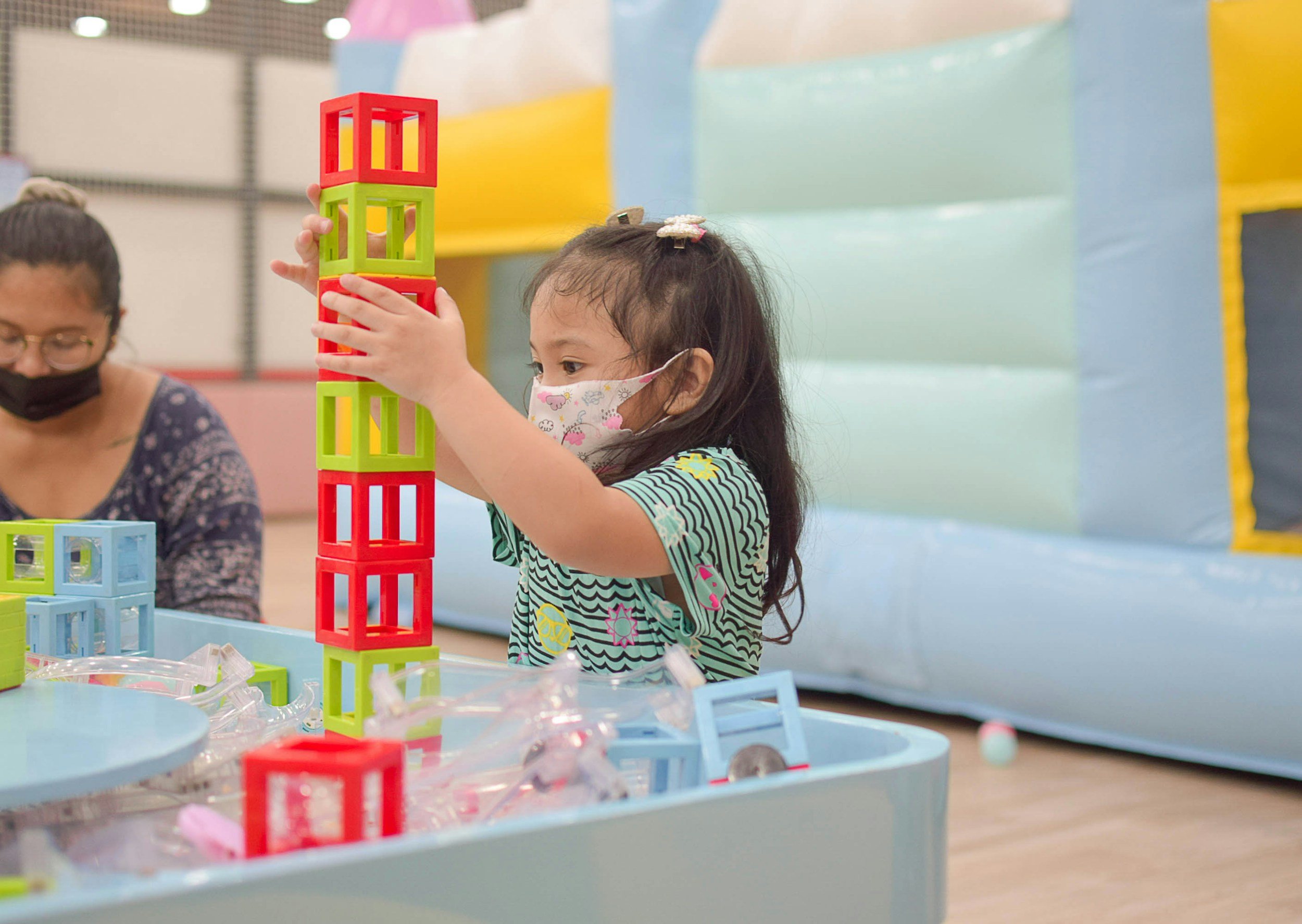 A young girl wearing a striped shirt and a face mask, stacking colorful plastic cubes in an indoor play area.