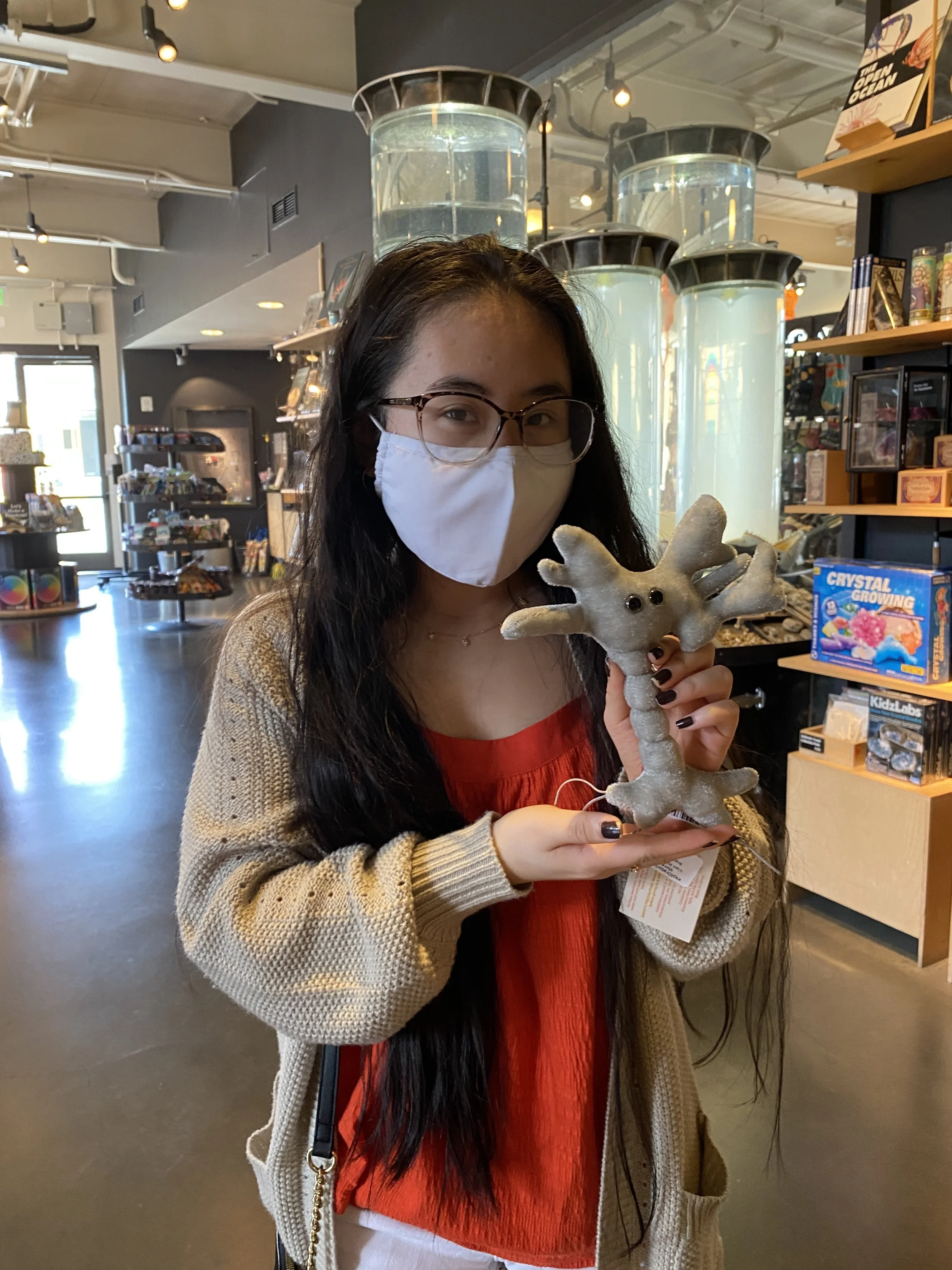Naomi wearing glasses and a face mask inside the Exploratorium, holding a gray neuron plushie.