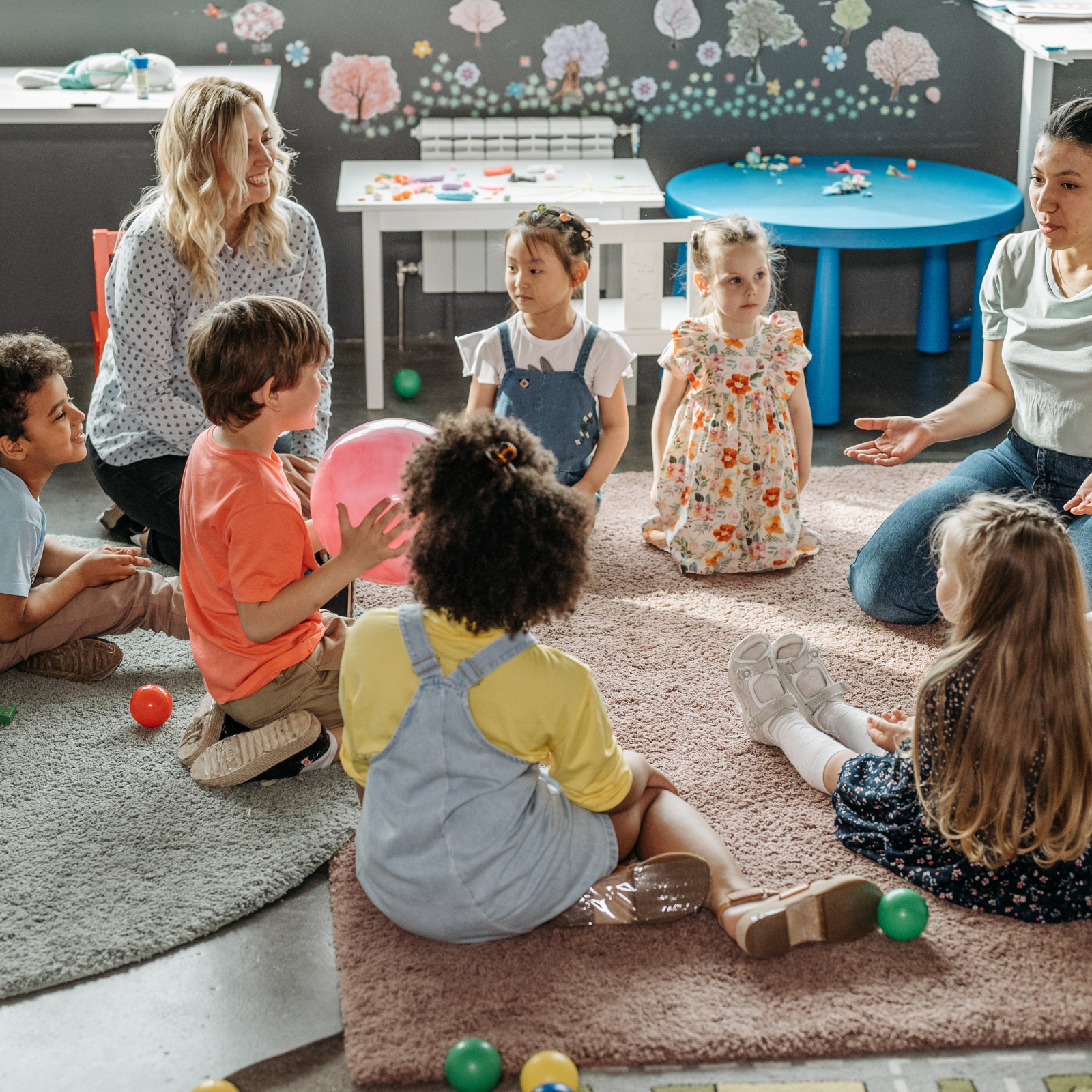 A group of young children and two adult women sitting on a beige carpet in a circle, engaging in a group activity.