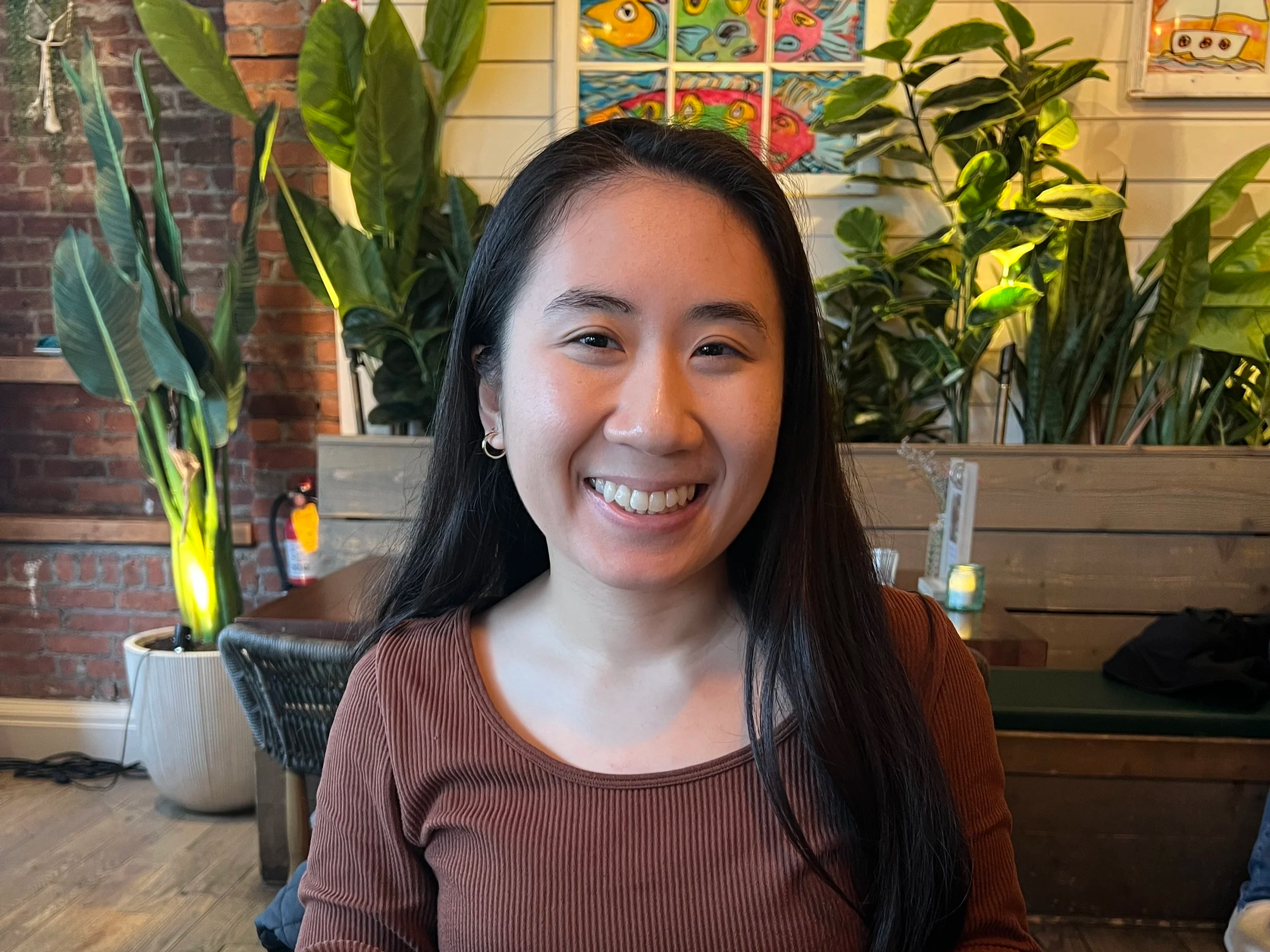 Naomi smiling in a brown top, seated indoors with large green plants and colorful artwork on the wall behind her.