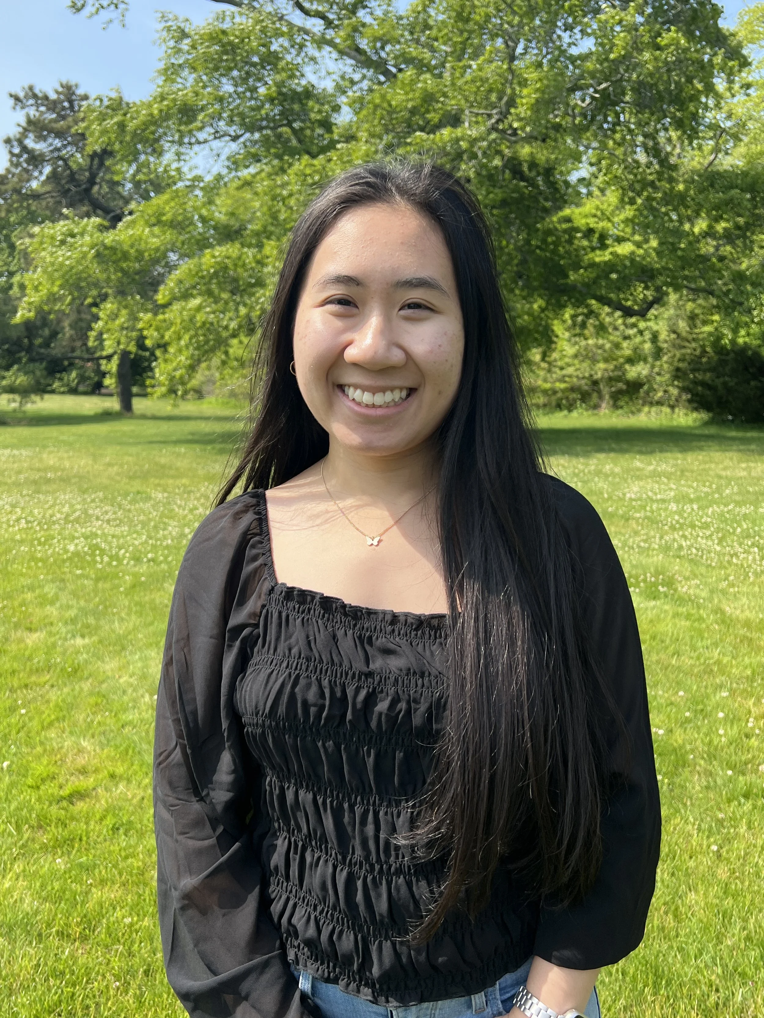 Naomi smiling, wearing a black top, standing in a park with green grass, trees, and blue sky in the background.