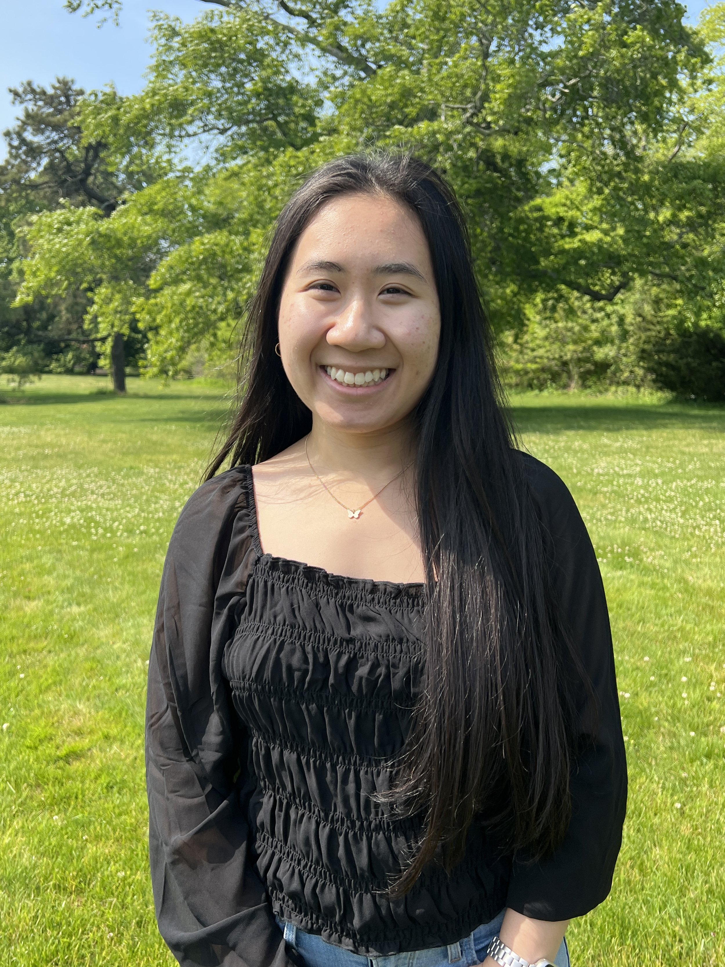 Naomi smiling, wearing a black top, standing in a park with green grass, trees, and blue sky in the background.