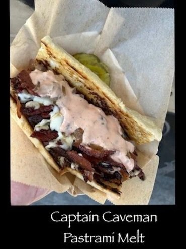 A sandwich in a paper wrapper containing pastrami, melted cheese, and sauce, topped with a pickle slice.
