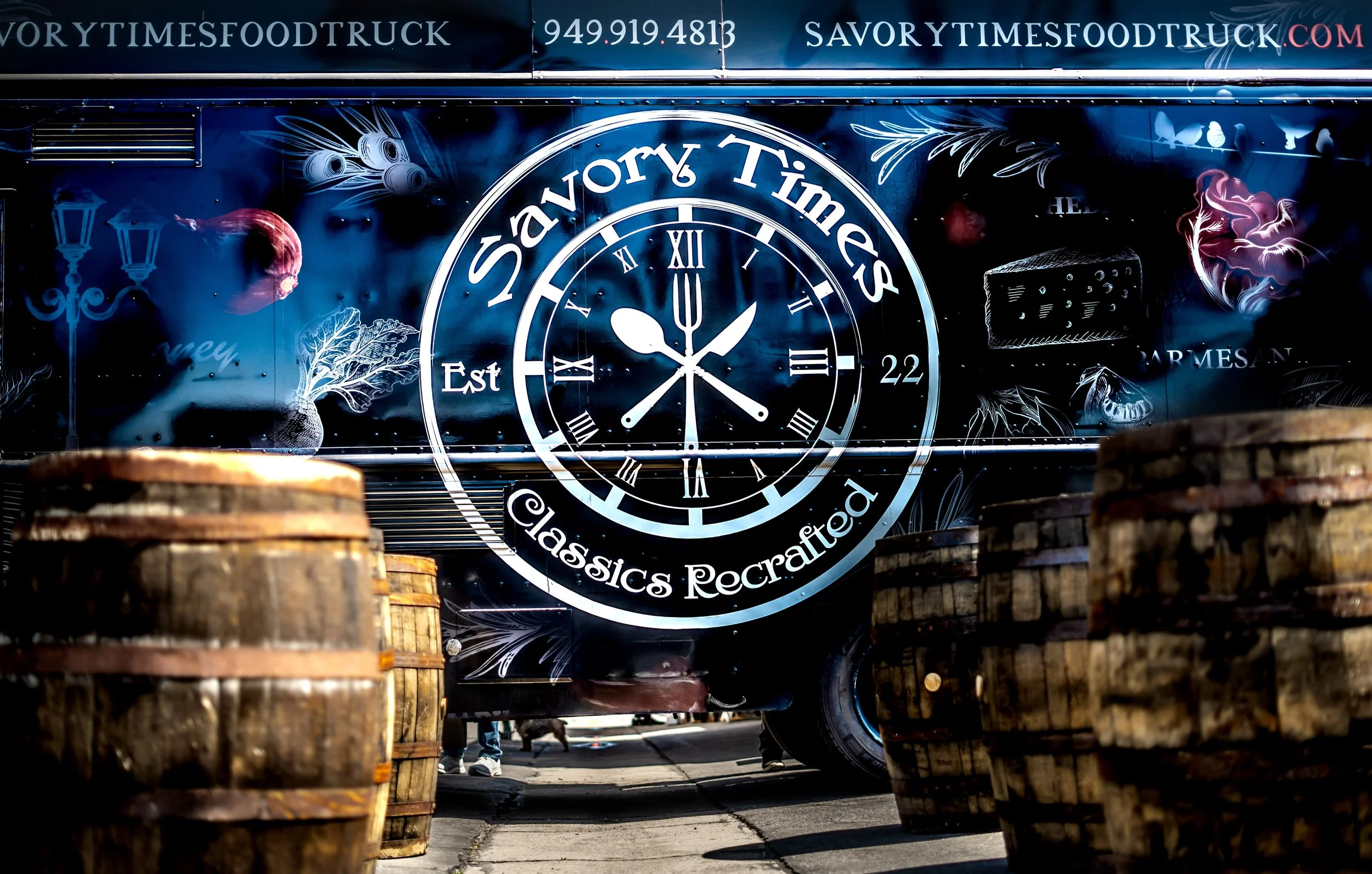 A food truck decorated with the words 'Savory Times Food Truck' and a clock design, parked behind wooden barrels on a street.