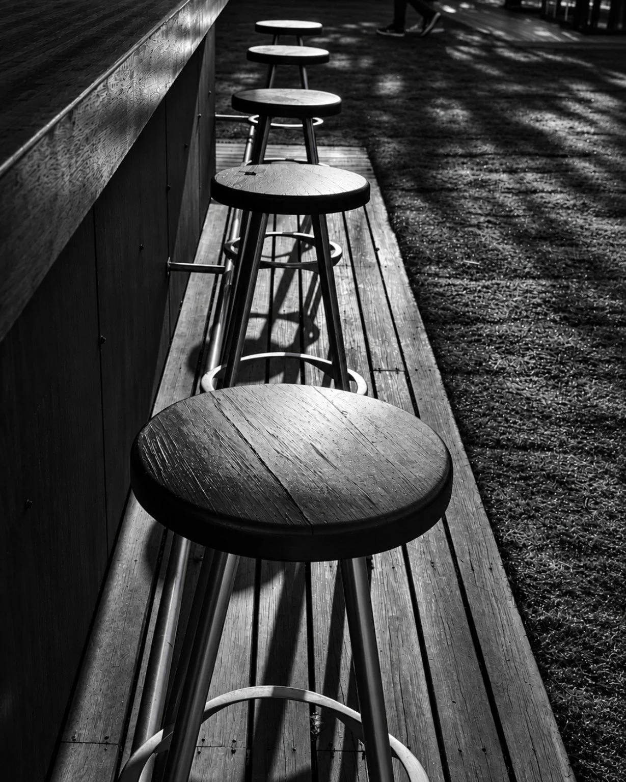 Black-and-white photograph of wooden bar stools receding beside an outdoor counter in patterned sunlight.