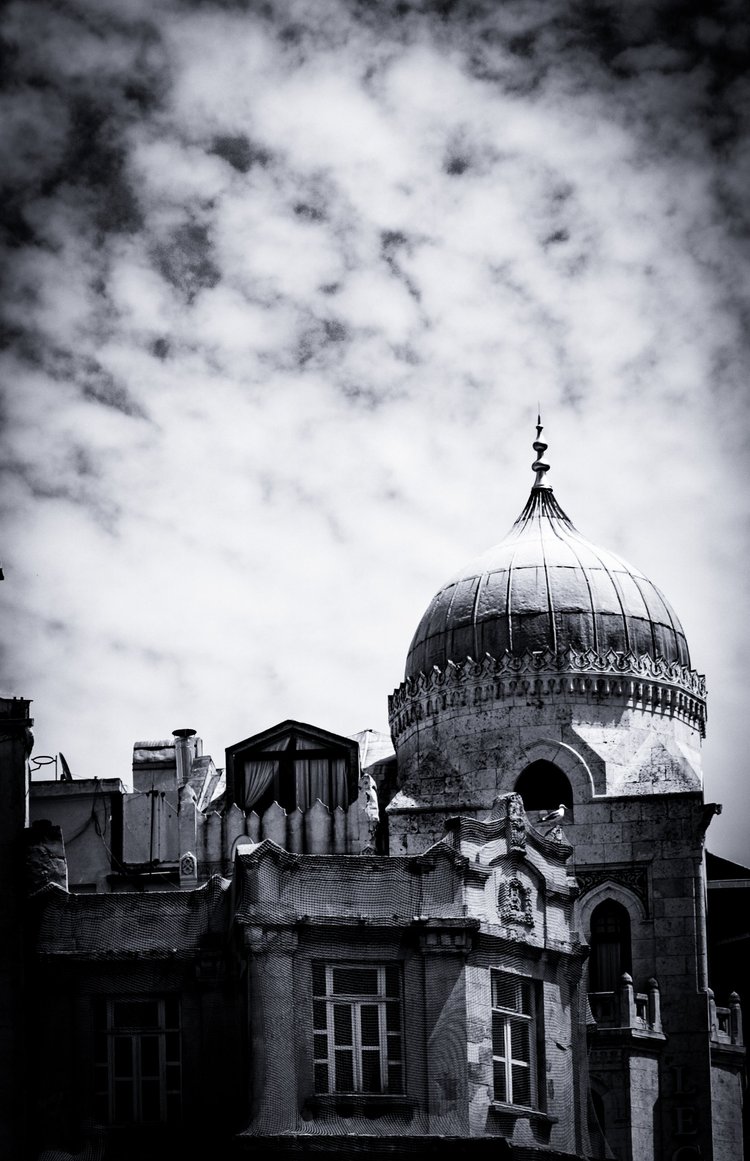 Black-and-white photograph of a weathered ornate building with a domed turret beneath textured clouds.