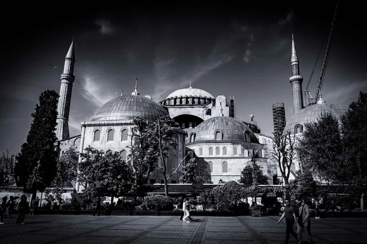 Black-and-white view of a monumental domed mosque and its minarets beyond a tree-lined public square.