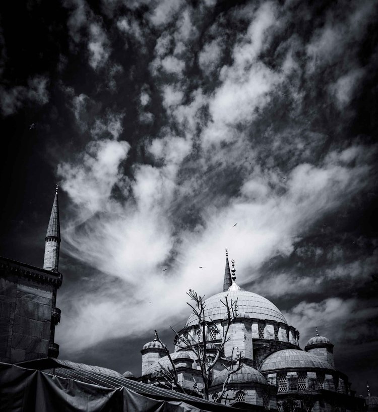 A mosque dome and minaret beneath sweeping, high-contrast clouds in a black-and-white sky.