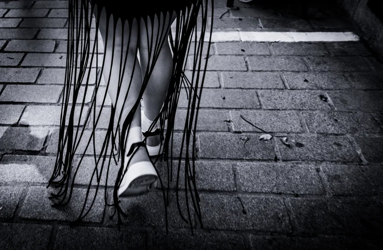 Black-and-white close-up of legs in sandals beneath long clothing fringe on a paved sidewalk.
