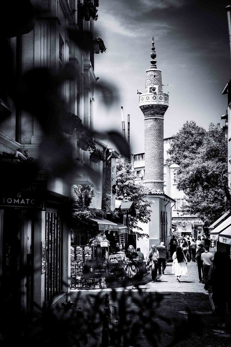 A minaret rises above a narrow shopping street framed by buildings and leafy shadows, in black and white.