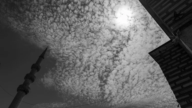 Black-and-white upward view of a minaret and roof edges framing a sky covered in rippling clouds.