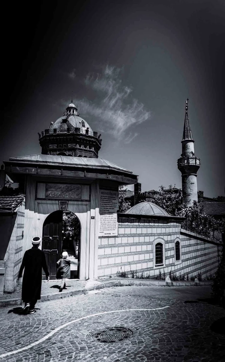 A person approaches an ornate gateway beside a stone wall, with a dome and minaret beyond, in black and white.