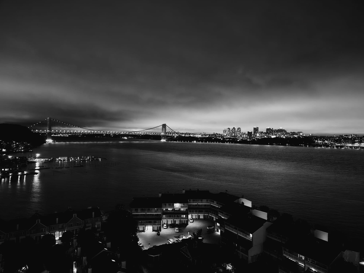 Black-and-white nighttime panorama of an illuminated bridge crossing a broad river beneath a cloudy sky.
