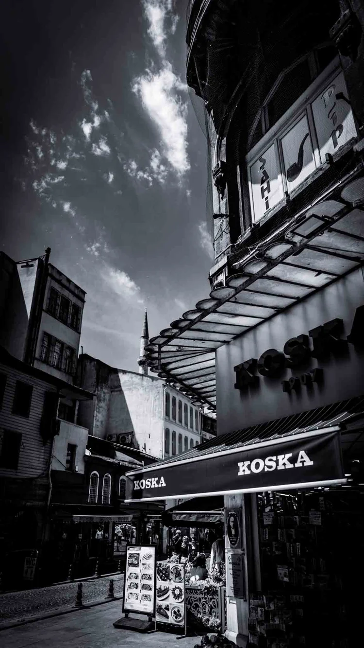 Black-and-white upward view along a shopping street, with a shop awning and glass canopy beneath tall buildings.