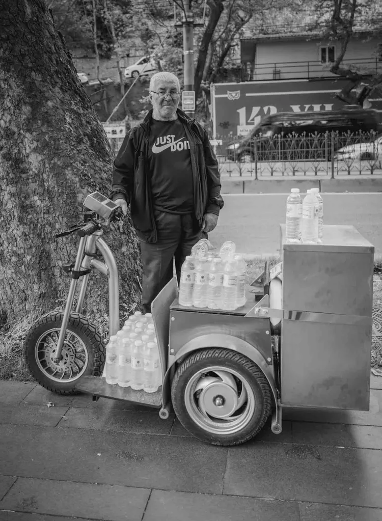 Black-and-white portrait of a man standing beside a cart carrying bottled water on a city sidewalk.