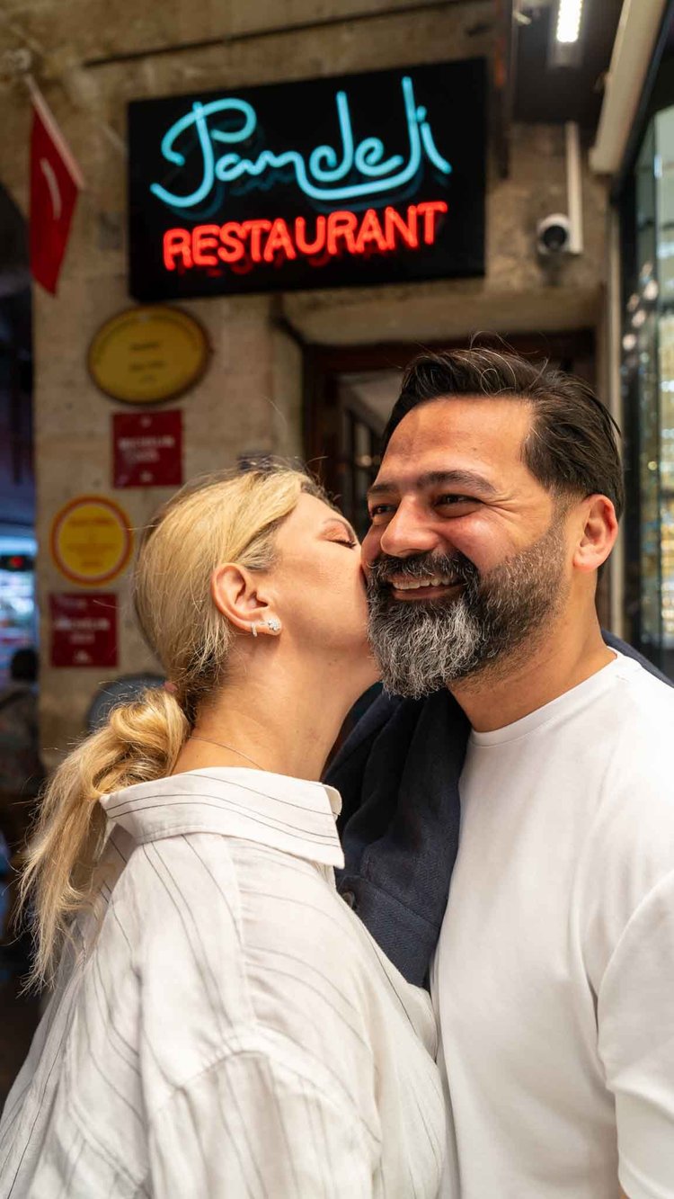 A woman kisses a smiling man on the cheek outside a restaurant entrance.