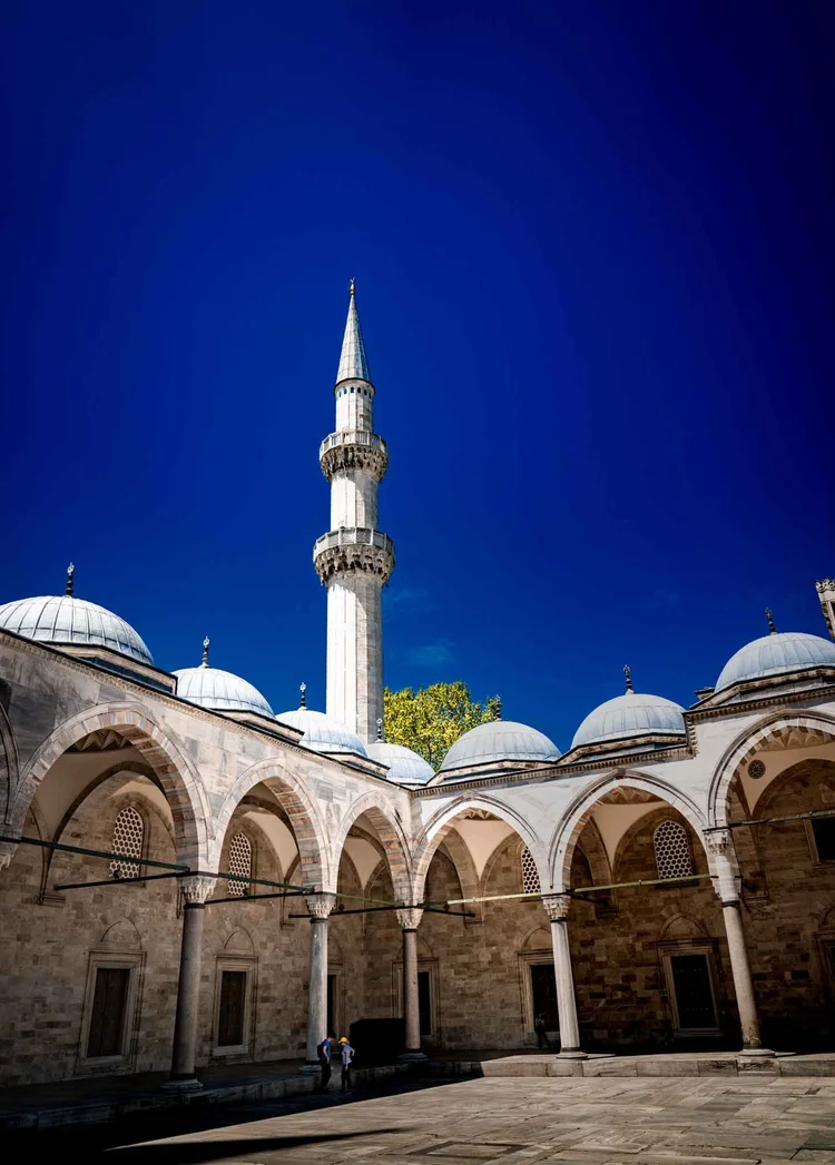 A mosque courtyard with stone arches, domes, and a slender minaret against a deep blue sky.