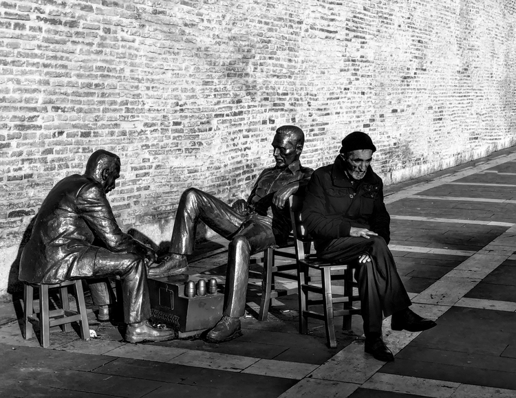 A man sits beside a sculptural group of seated figures against a long brick wall, in black and white.