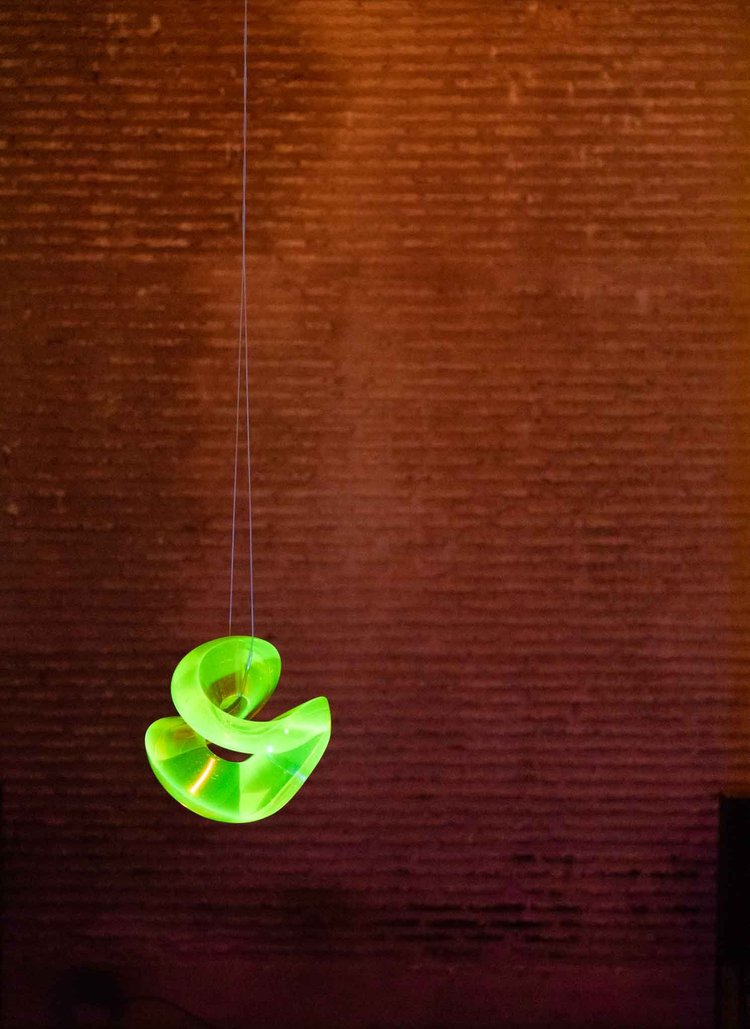 A glowing green abstract sculpture suspended against an orange-lit brick wall.