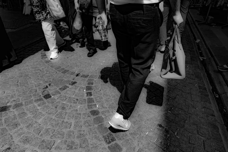 Black-and-white close-up of pedestrians’ legs, shoes, and shadows crossing a cobbled shopping street.
