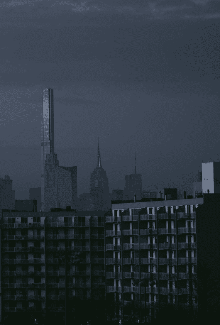 A distant skyline with a slender skyscraper rises beyond a dark foreground building beneath a blue-gray sky.