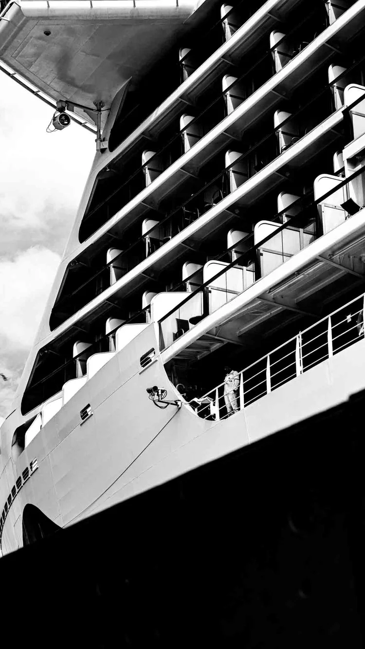 Black-and-white low-angle view of a large ship, showing stacked decks, railings, and lifeboats.