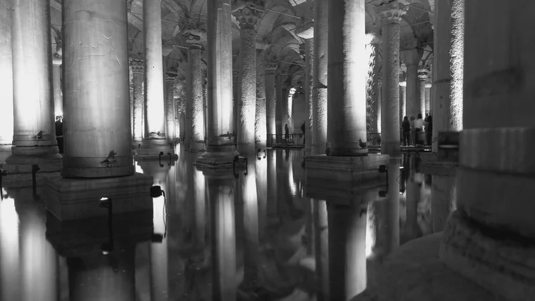 Illuminated stone columns reflected in still water inside a vaulted underground cistern, in black and white.