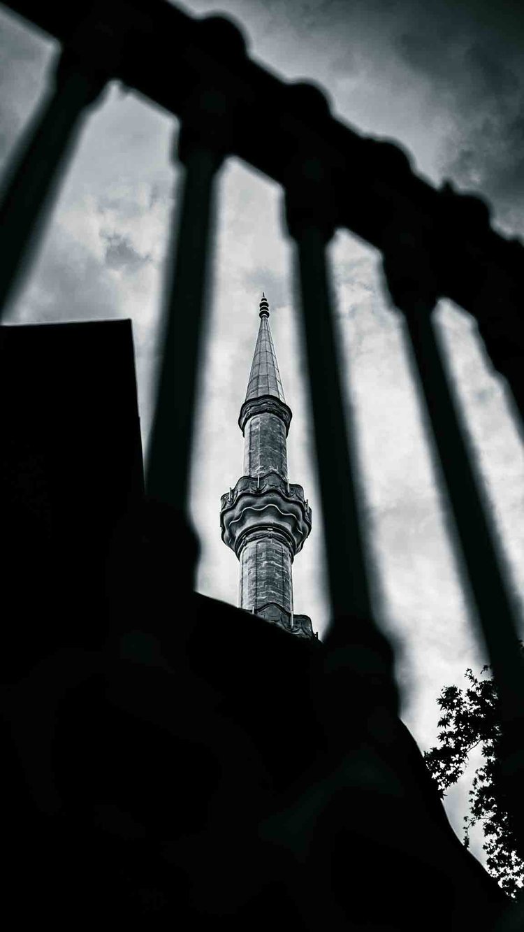 A slender minaret seen through dark foreground railings beneath a cloudy sky.