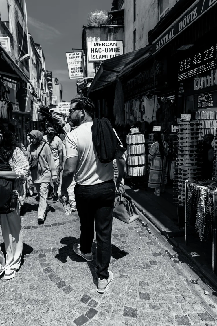 Black-and-white street photograph of a man walking through a narrow lane lined with shop displays.