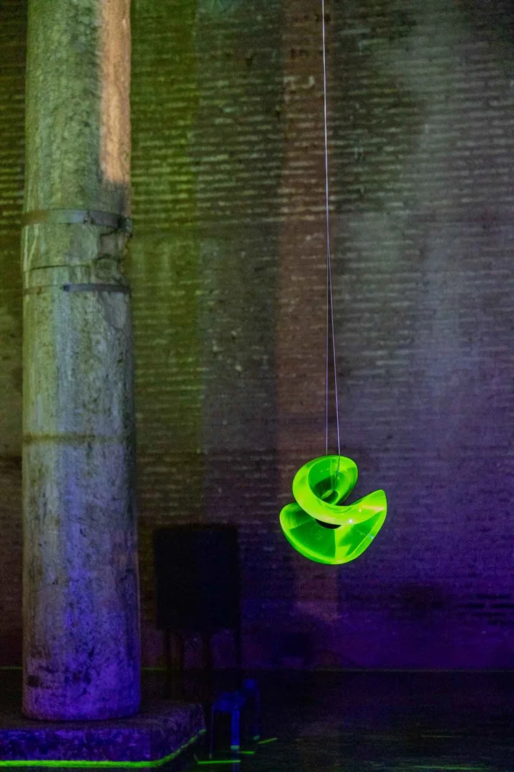 A luminous green sculpture hangs beside a stone column washed in purple and green light.