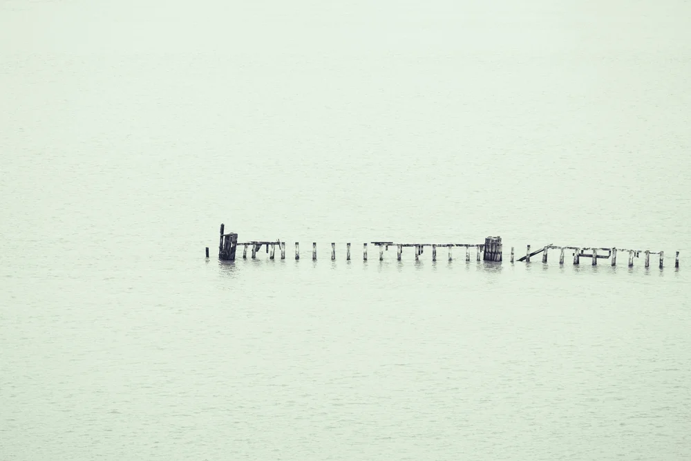 A distant, weathered pier stretches across pale, still water in a minimalist, misty landscape.