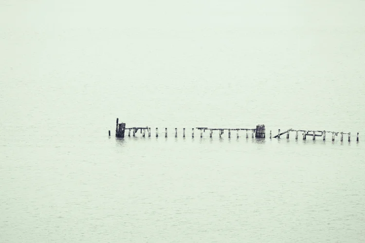 A distant, weathered pier stretches across pale, still water in a minimalist, misty landscape.