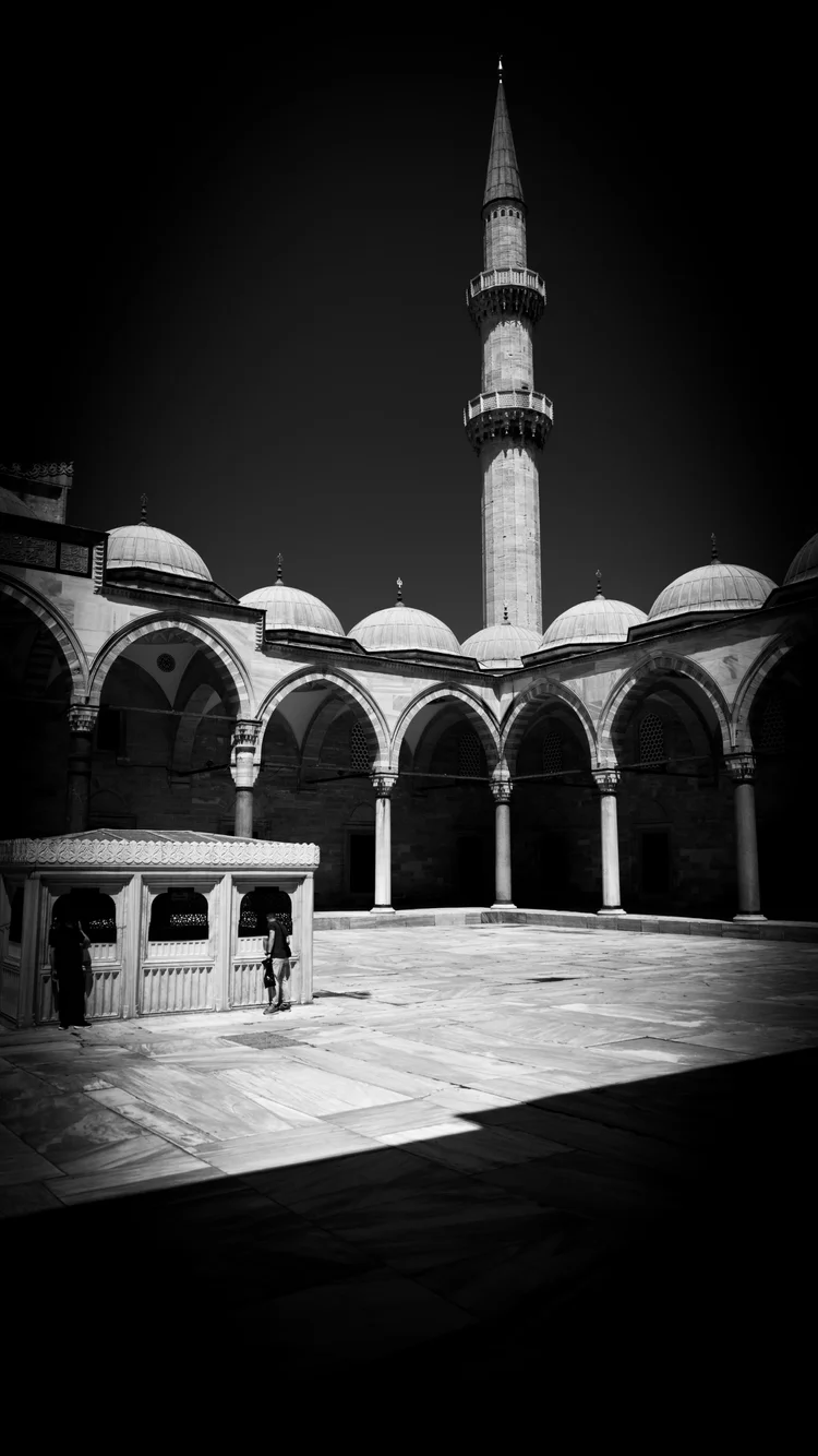 Black-and-white photograph of a sunlit mosque courtyard with repeating arches and a tall minaret.