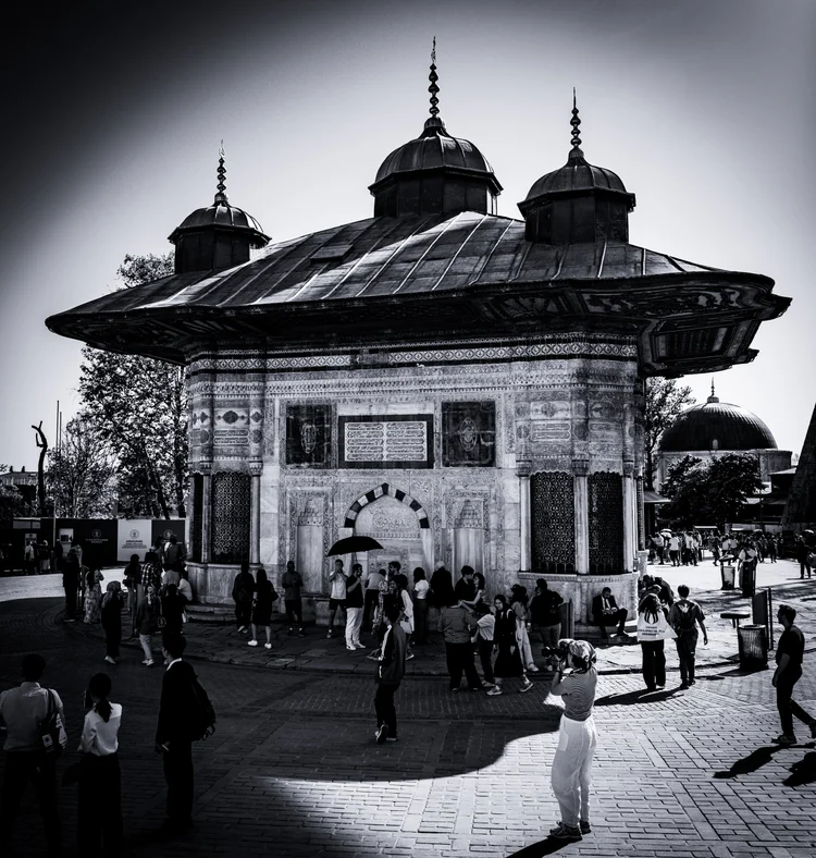 Black-and-white photograph of an ornate fountain pavilion surrounded by people in a sunlit square.