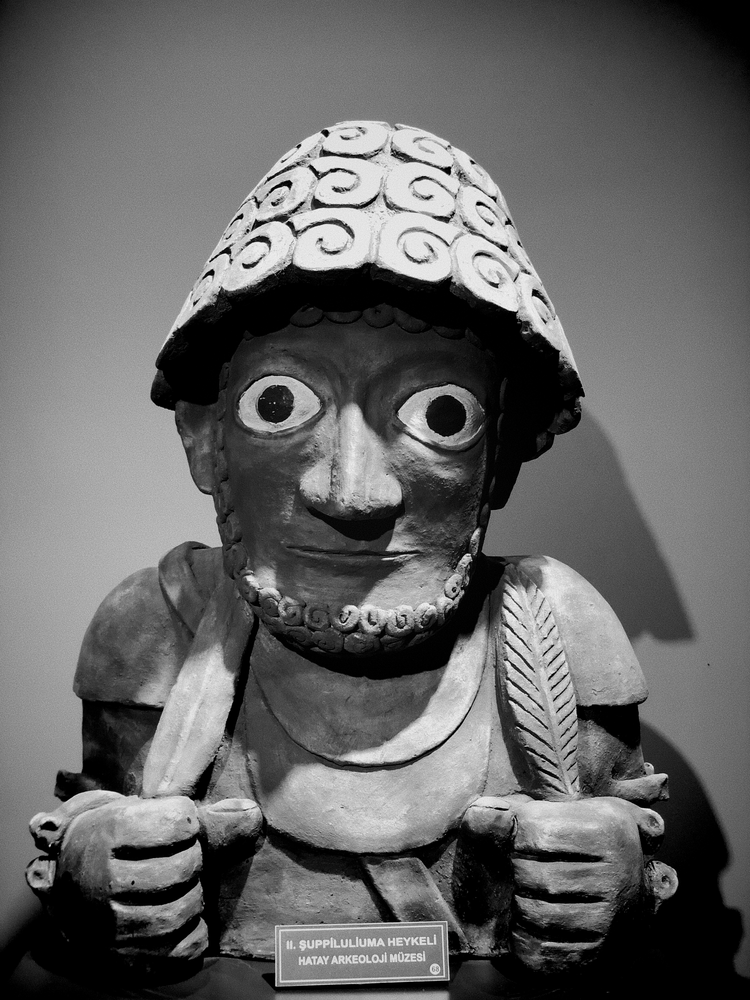 Black-and-white close-up of a sculpted figure with wide eyes, an ornate helmet, and hands raised near its chest.