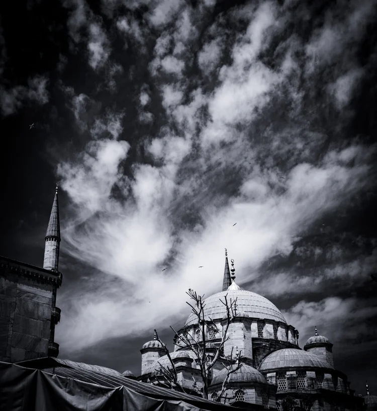 A mosque dome and minaret beneath sweeping, high-contrast clouds in a black-and-white sky.
