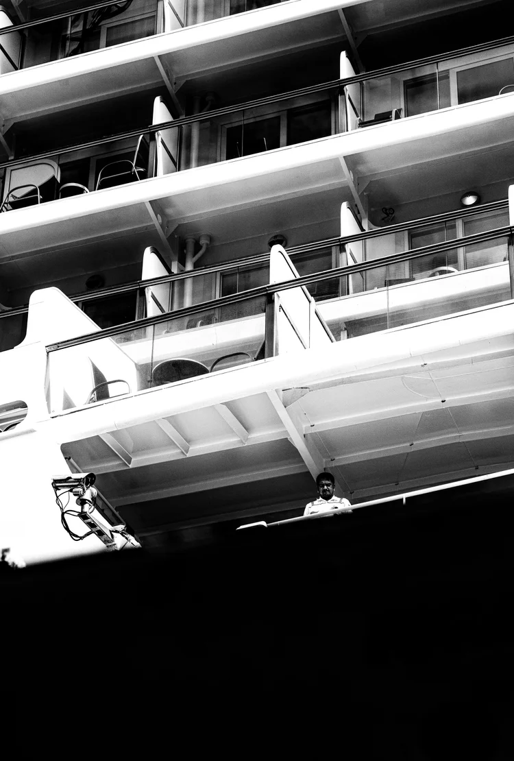 Black-and-white close-up of a ship’s white lifeboat and stacked balcony railings against deep shadow.