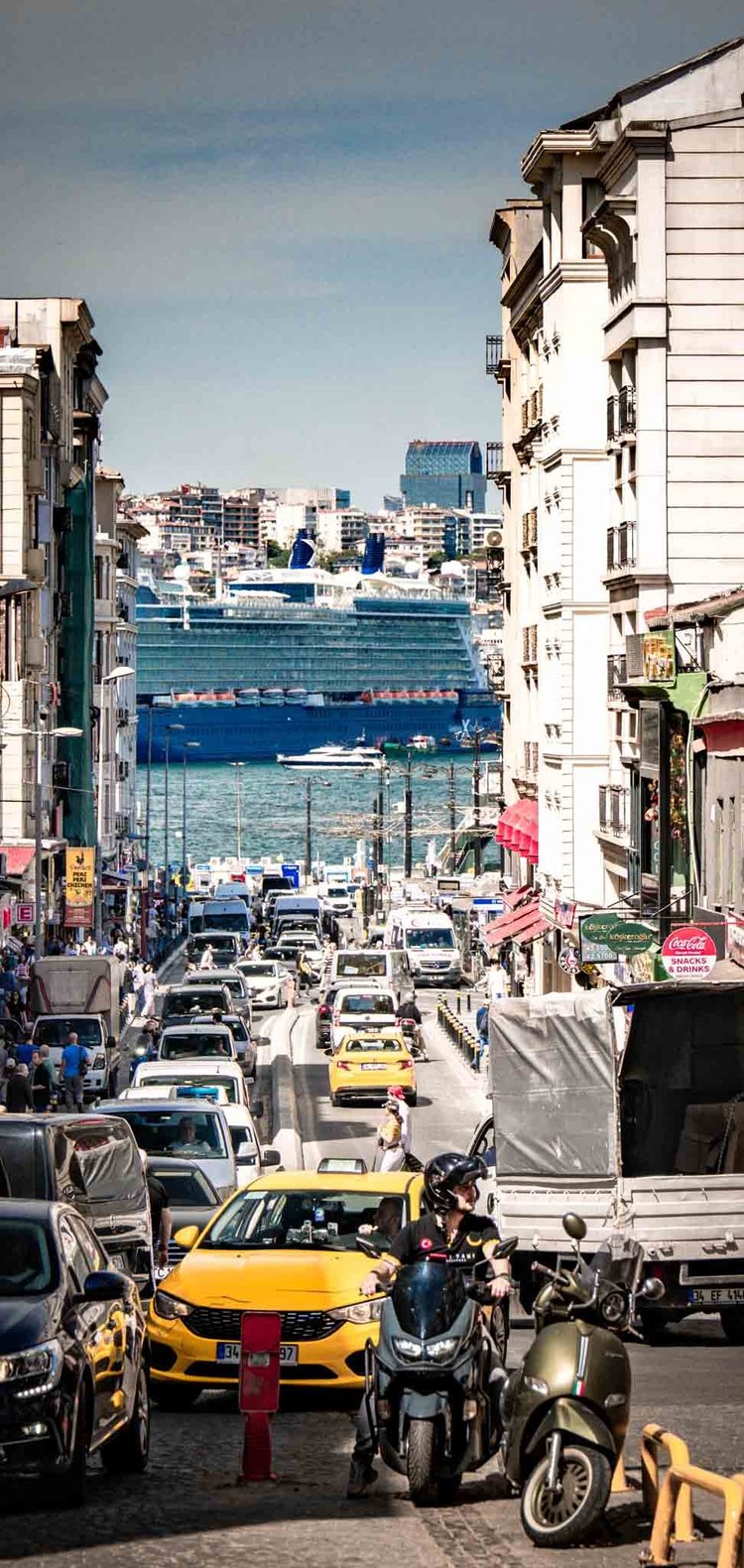 A busy street with yellow taxis leads downhill toward blue water and a large passenger ship.
