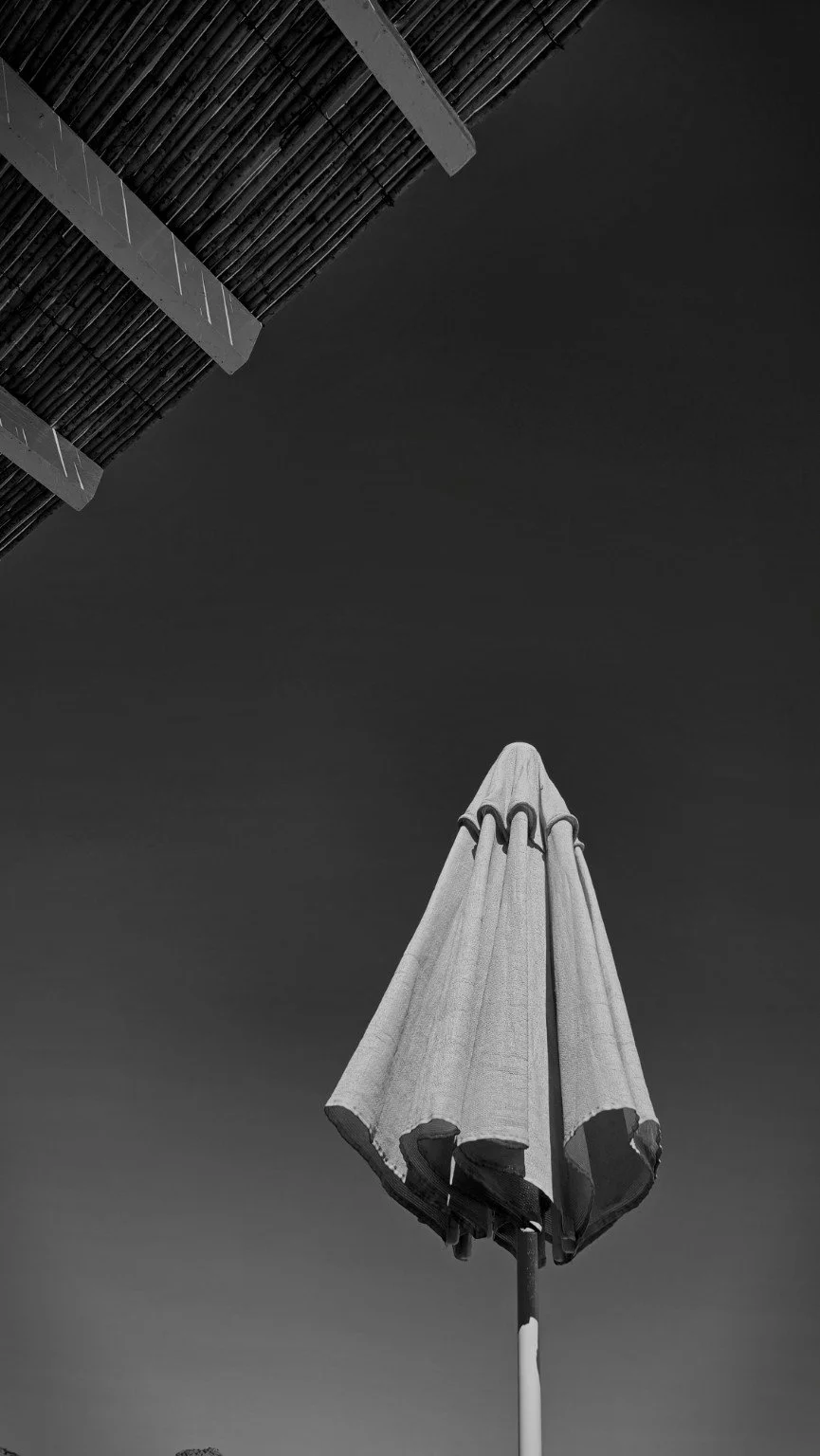 Black-and-white upward view of a closed patio umbrella beneath the diagonal edge of a slatted canopy.
