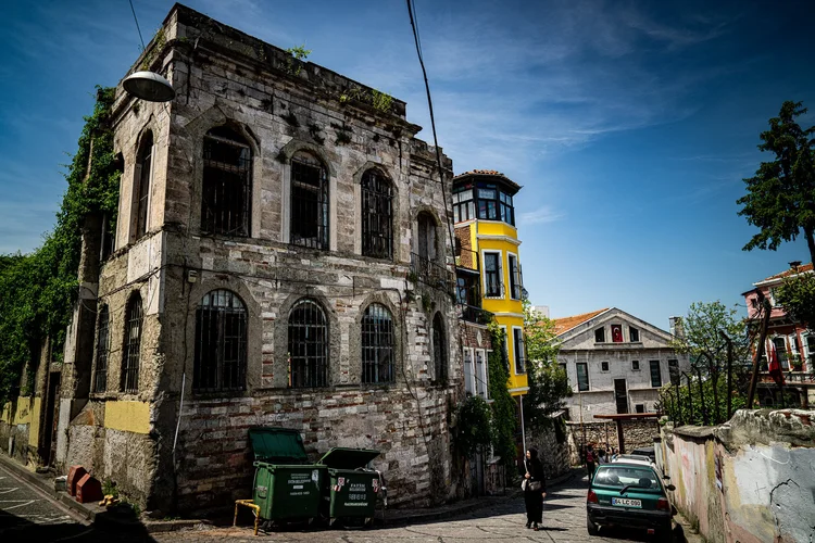 A weathered stone building with empty windows stands beside a narrow street under a blue sky.