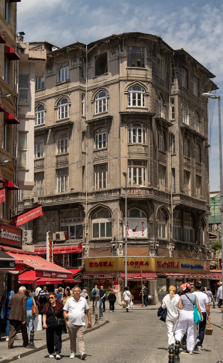 Pedestrians walk past shops beneath an ornate corner building with rows of projecting windows.