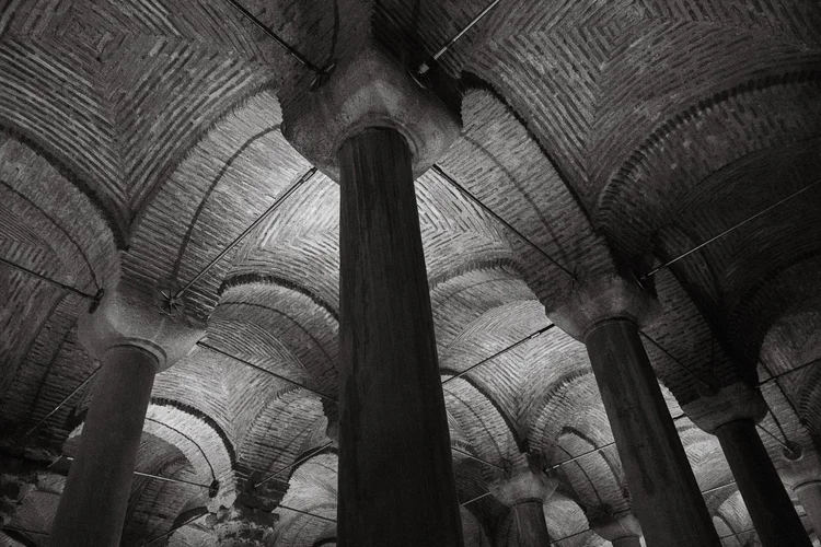 Black-and-white upward view of stone columns supporting a vaulted brick ceiling.