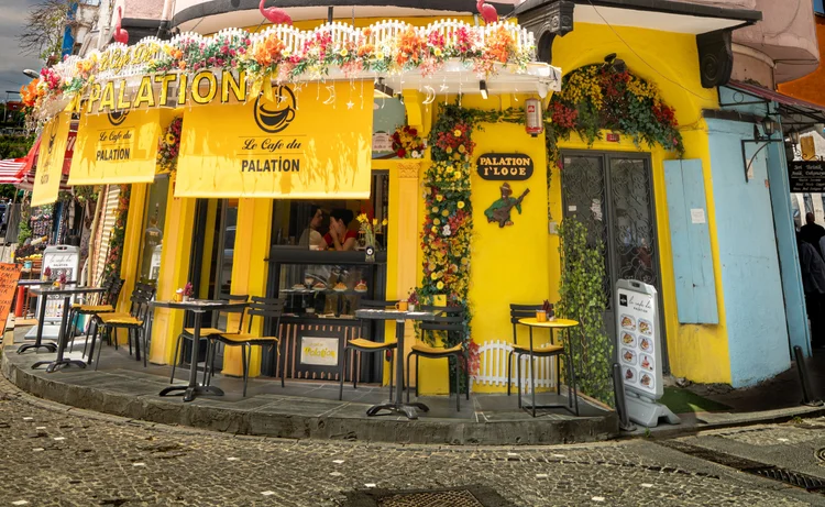 A bright yellow corner cafe decorated with flowers, with small tables along a cobbled street.