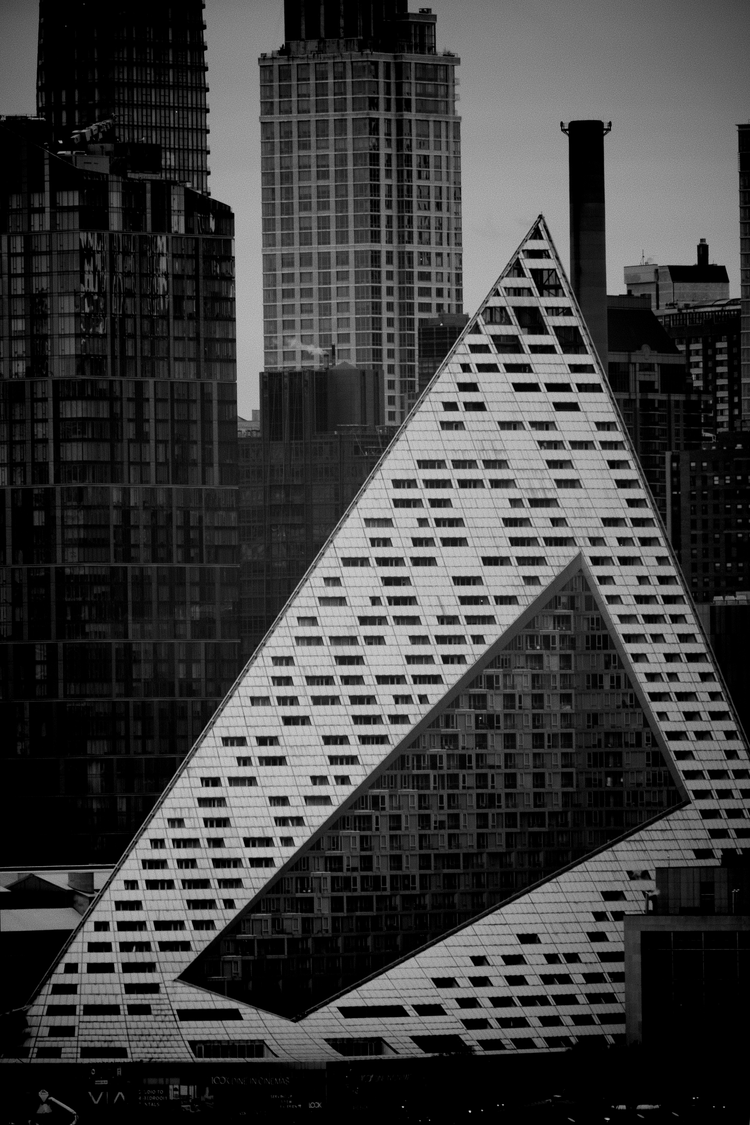 Black-and-white view of a triangular modern building with a patterned facade against taller city towers.