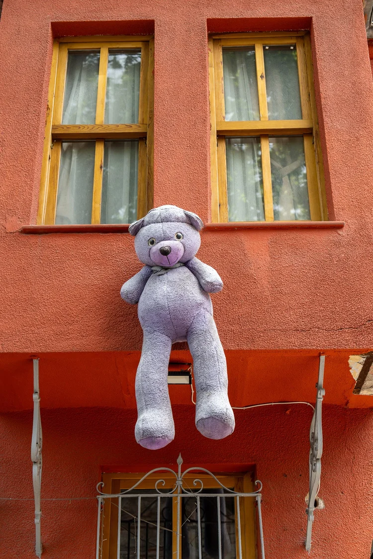 A pink teddy bear hangs in front of an orange building with yellow-framed windows.