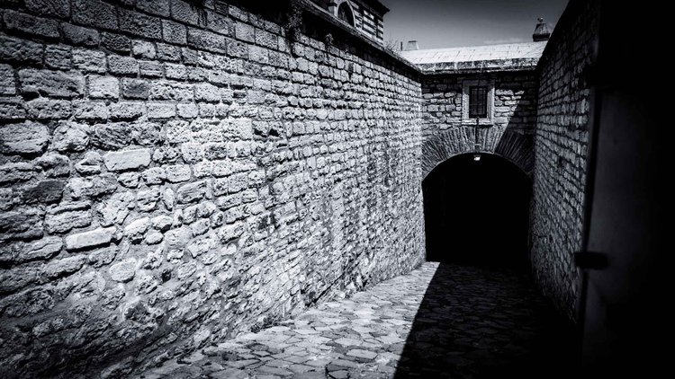 Black-and-white view of a narrow stone passage leading down to a dark arched doorway.