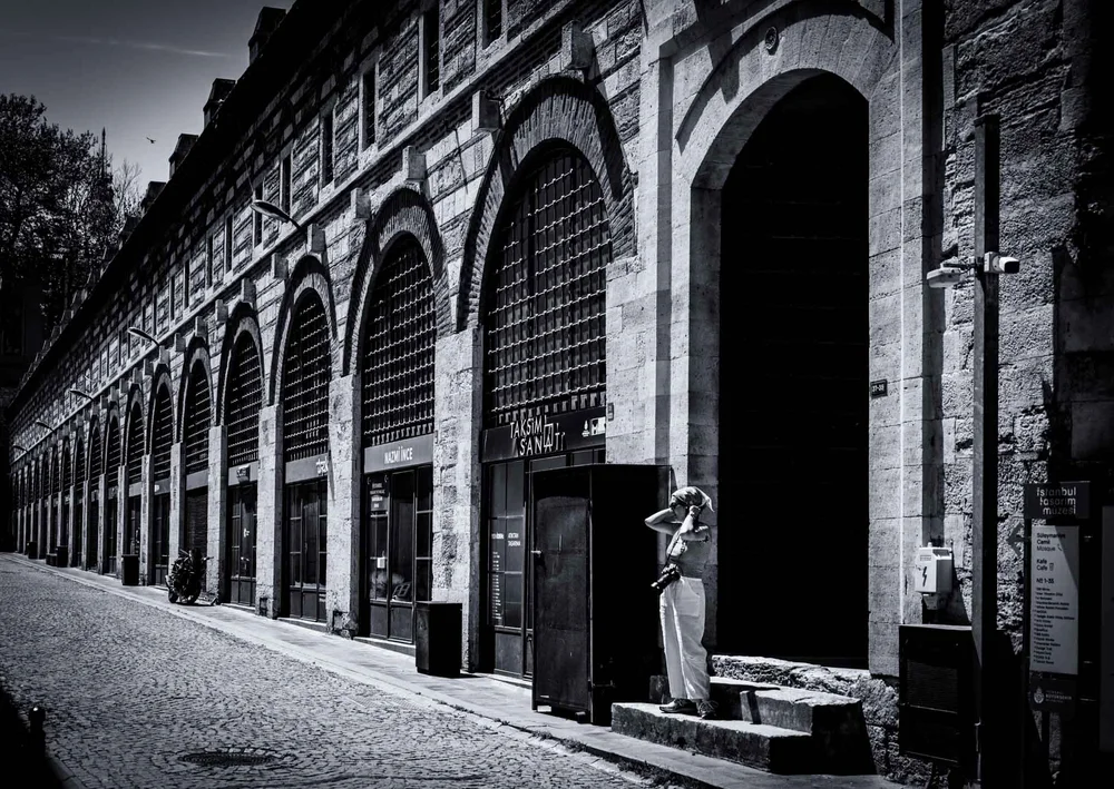 Black-and-white view along a cobbled street lined with tall stone arches, with a person standing in a doorway.