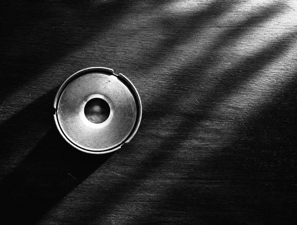 Black-and-white overhead view of a round metal ashtray on a wooden table crossed by light and shadow.