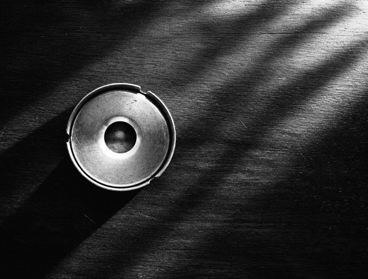 Black-and-white overhead view of a round metal ashtray on a wooden table crossed by light and shadow.