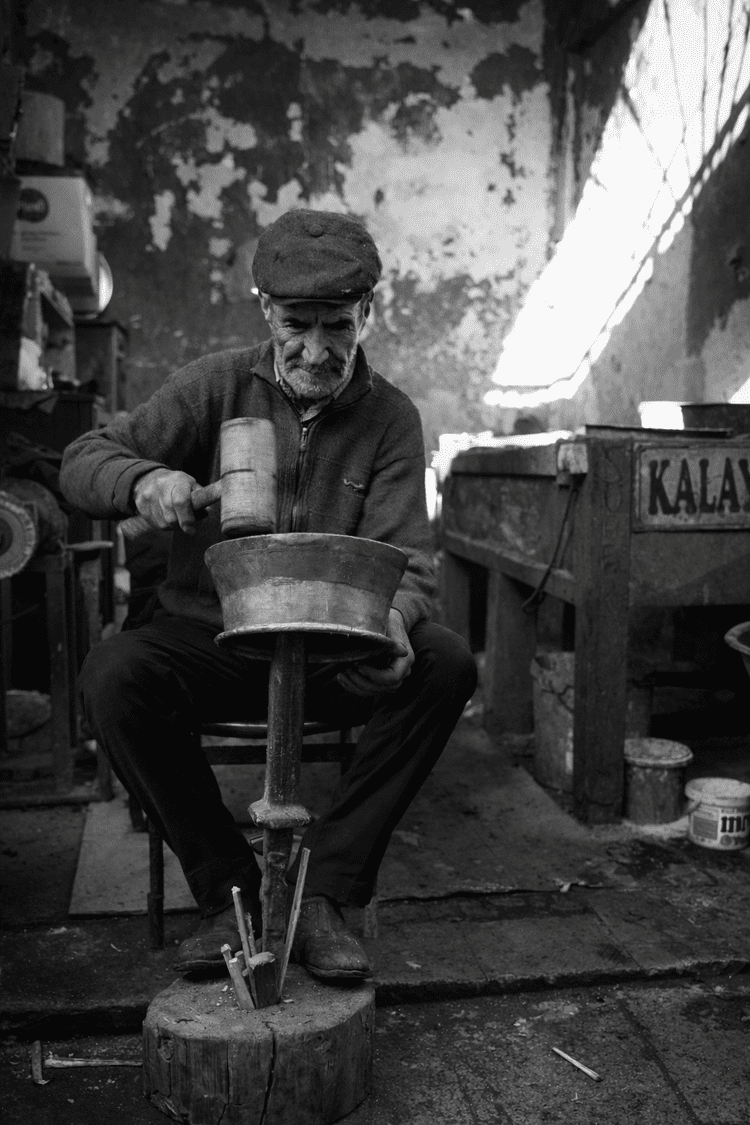 Black-and-white portrait of a seated metalworker shaping a bowl in a small workshop.